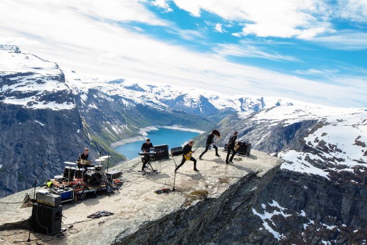 Norveçli Shining'i karla kaplı dağların önündeki kayalık zirvede performans sergilerken gösteren grup fotoğrafı