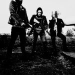 Deathcult'ın üç üyesini siyah kıyafetler ve metal aksesuarlarla açık arazide gösteren siyah beyaz grup fotoğrafı