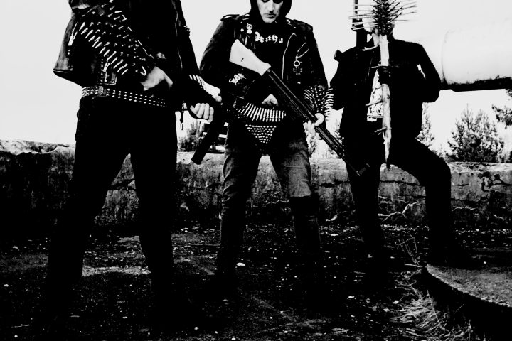 Deathcult'ın üç üyesini siyah kıyafetler ve metal aksesuarlarla açık arazide gösteren siyah beyaz grup fotoğrafı
