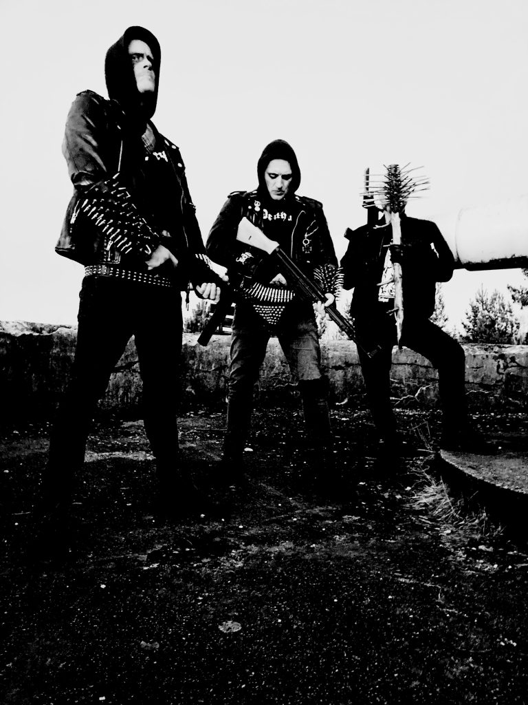 Deathcult'ın üç üyesini siyah kıyafetler ve metal aksesuarlarla açık arazide gösteren siyah beyaz grup fotoğrafı